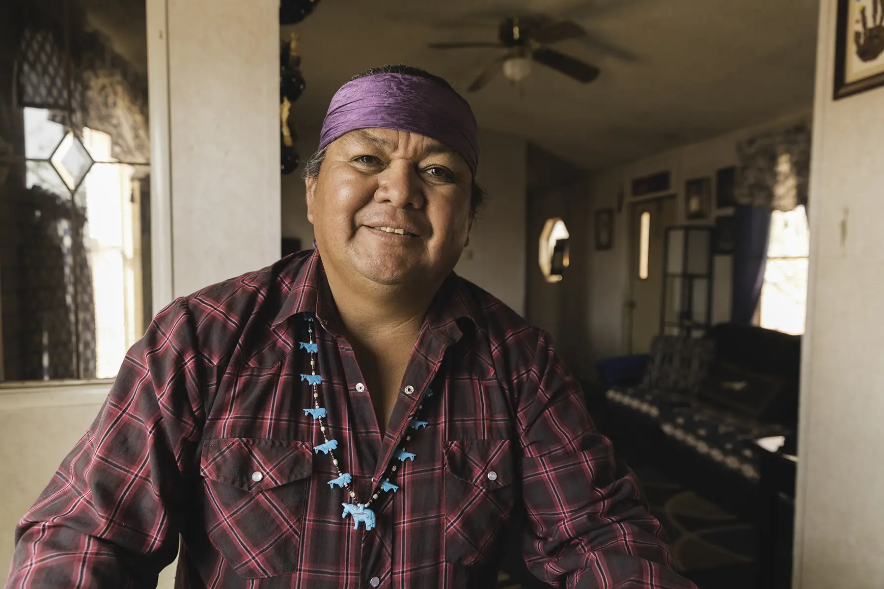 senior-in-living-room Man with purple headband, red striped shirt and necklace with turquoise horses standing in home.