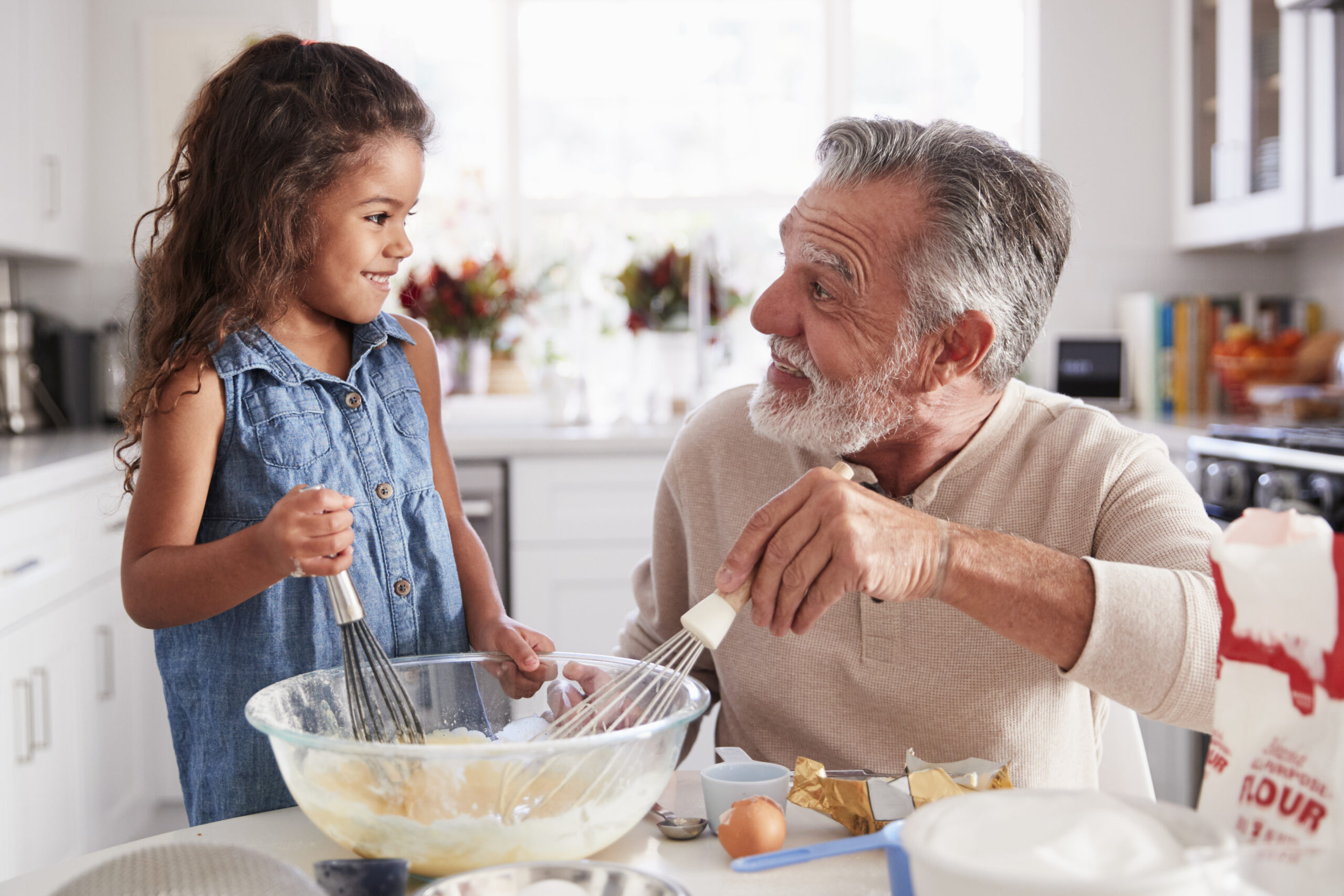 Young girl and grandfather making cake mixture at the kitchen table, smiling at each other