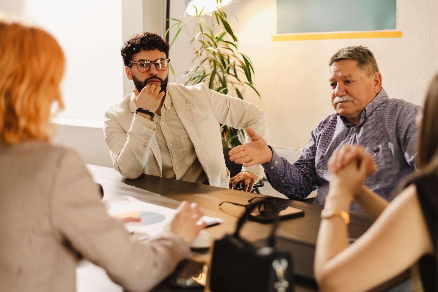 Four professionals engage in a lively discussion in a contemporary office. One man gestures while explaining, and the others listen attentively. Natural light fills the space.