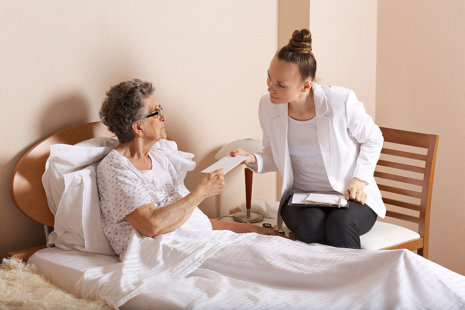 a woman hands an envelope to an older woman at her bedside