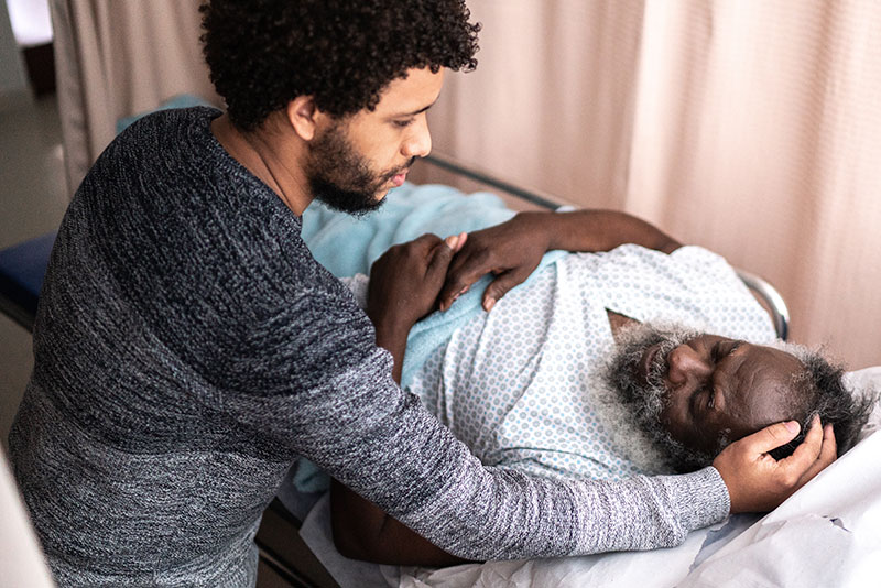 A man with a beard leaning over another man in a caring way . the other man is black and in a hospital bed.