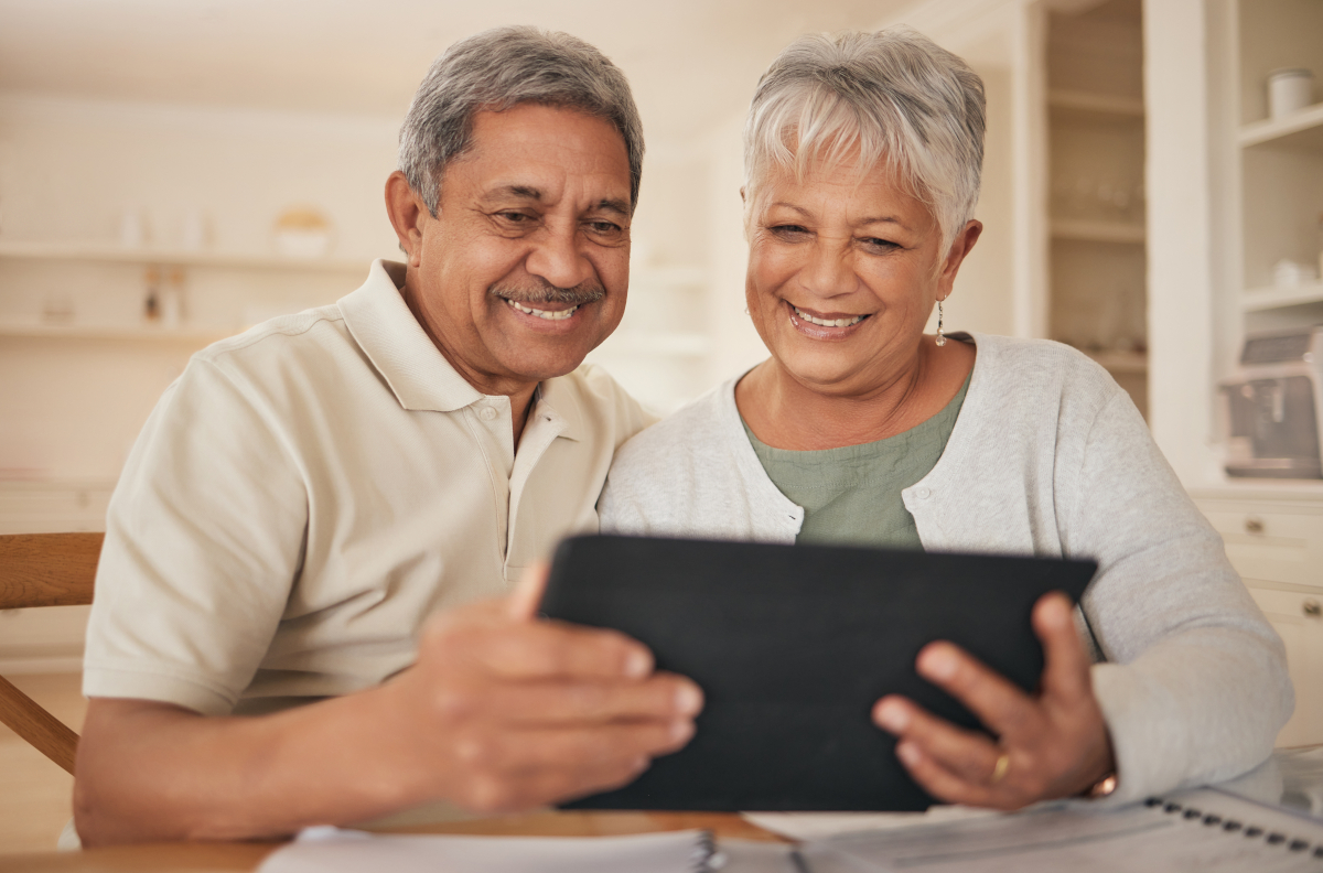 A senior hispanic couple looking at a tablet computer. The man on the left is in a cream colored shirth with a mustach. The Woman on the right has silver hair. Both are smiling.