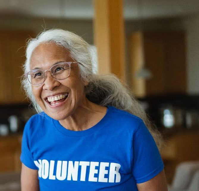 Smiling woman with gray hair and glasses wearing a blue tshirt with the word volunteer in white lettering across the front