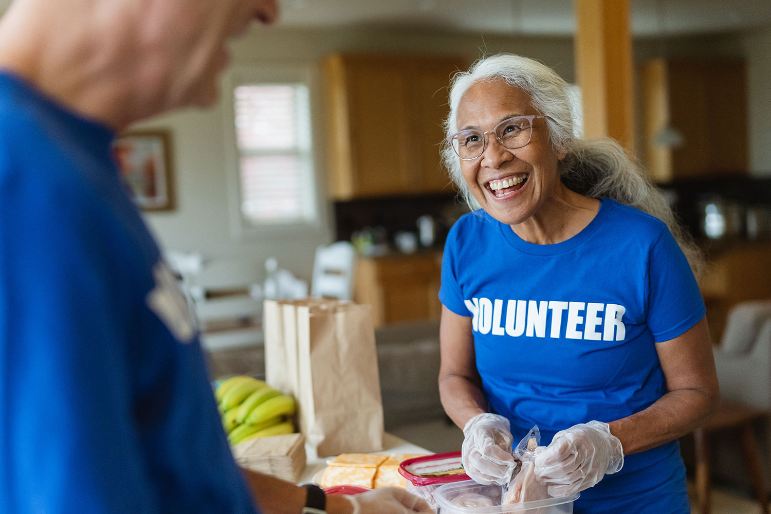 A woman with grey hair wearing glasses, she is smiling and wearing a blue t shirt with the word volunteer across the front.